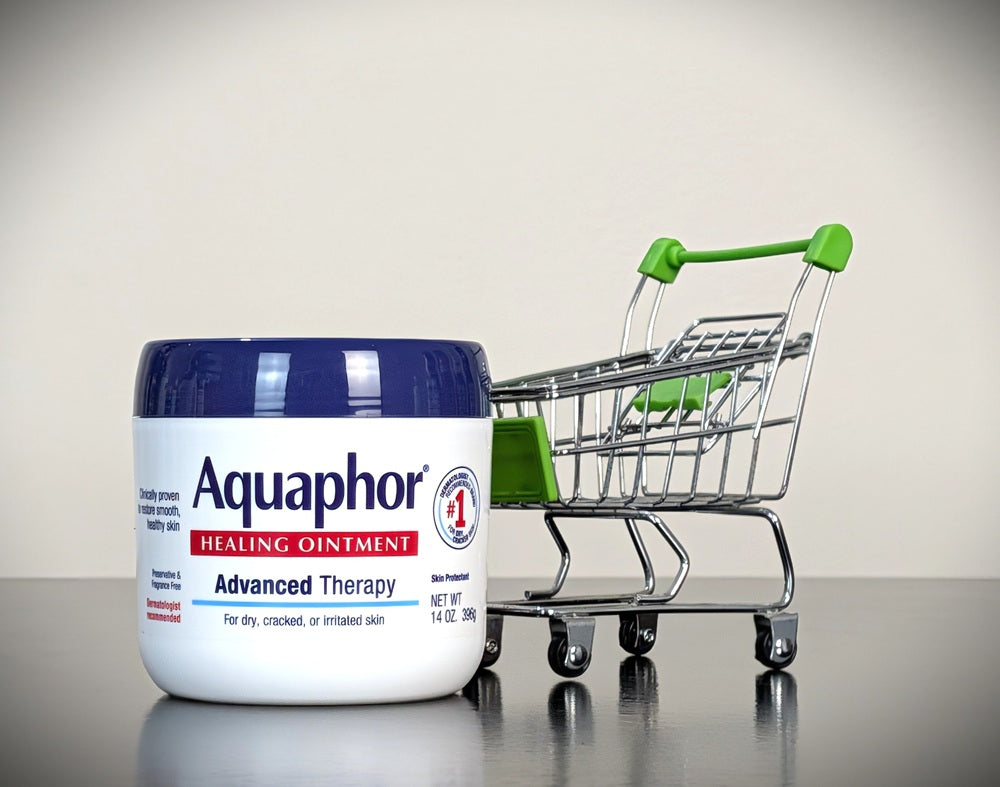 A white jar of Aquaphor Healing Ointment with a dark blue lid sits on a reflective surface next to a miniature metal shopping cart with a green handle.