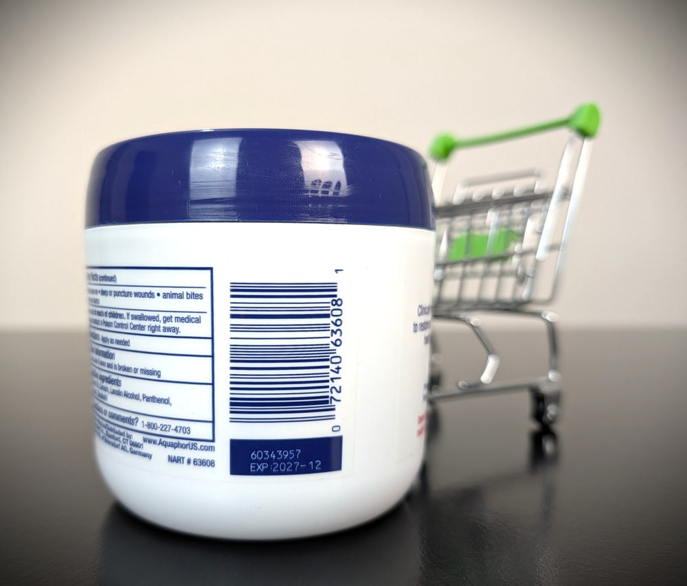 A white jar of Aquaphor Healing Ointment with a dark blue lid sits on a reflective surface next to a miniature metal shopping cart with a green handle.