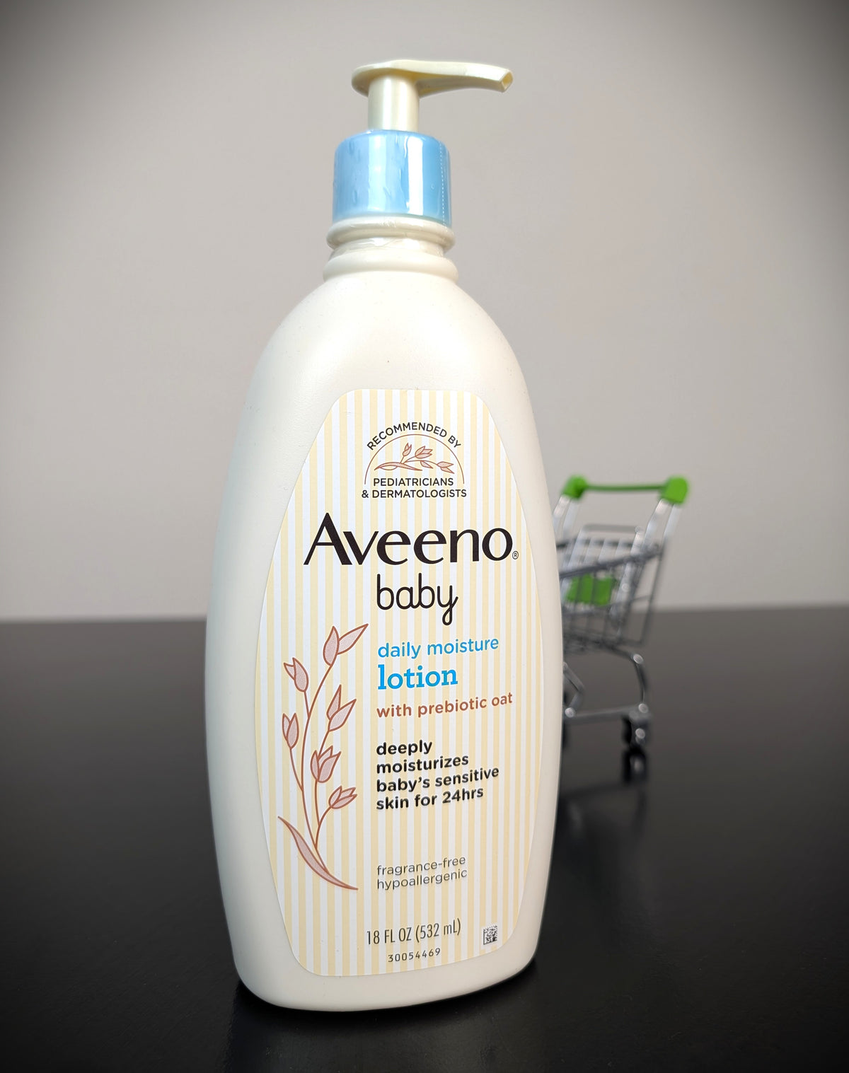 A bottle of Aveeno Baby Daily Moisture Lotion with a pump dispenser, standing on a dark surface against a light gray background. A miniature shopping cart is visible in the blurred background.