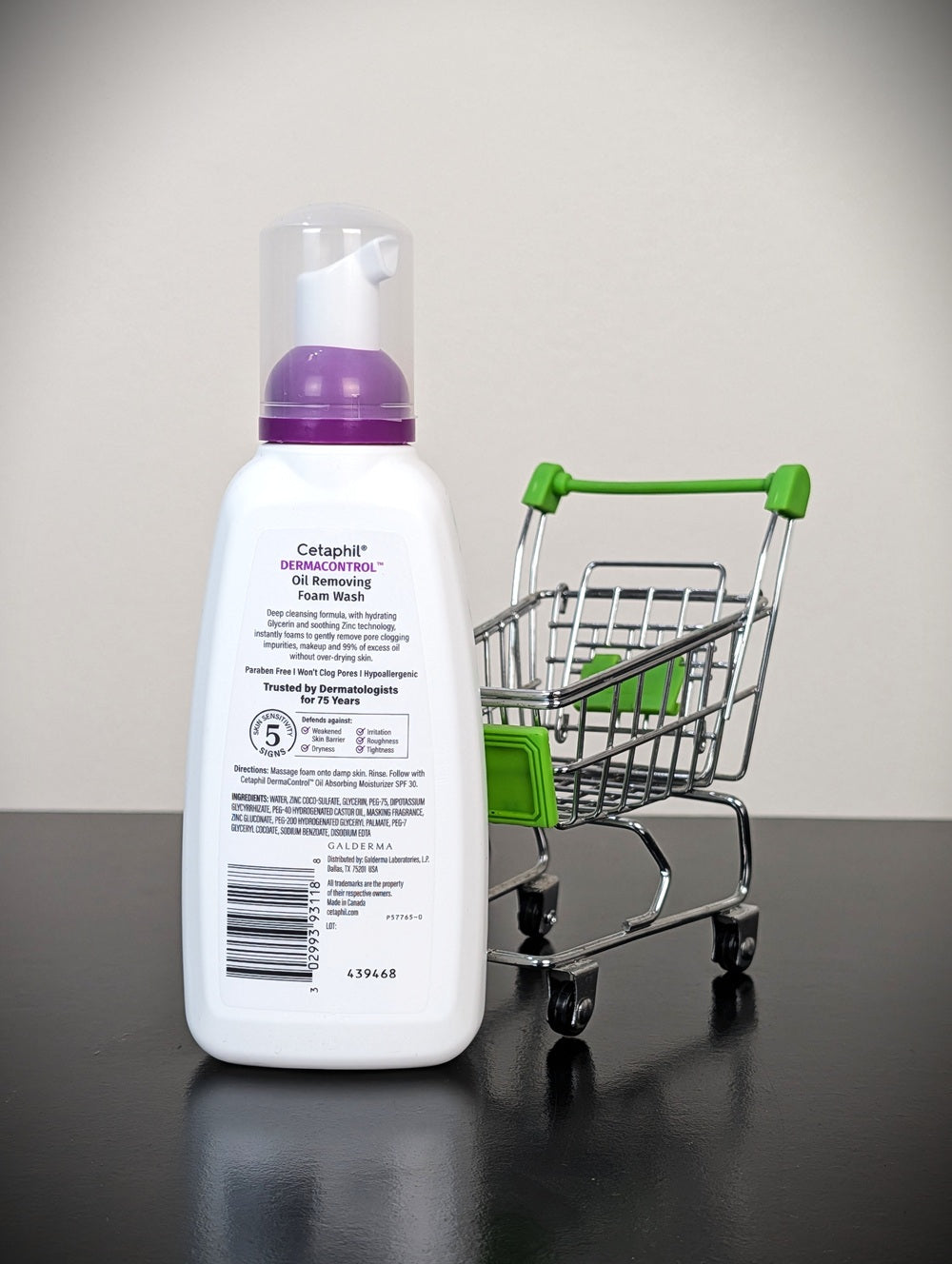 A bottle of Catapoint Depigmenting Foam Wash with a purple pump, standing next to a miniature metal shopping cart with a green handle on a dark reflective surface against a light gray background.