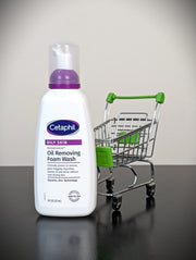 A bottle of Cetaphil Oily Skin Oil Removing Foam Wash stands on a dark reflective surface next to a small metal shopping cart with green handles and seat.