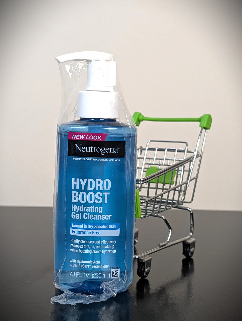 A bottle of Neutrogena Hydro Boost Hydrating Gel Cleanser in blue packaging, wrapped in a clear plastic protective sleeve, standing on a dark surface next to a small metal shopping cart with green handles.