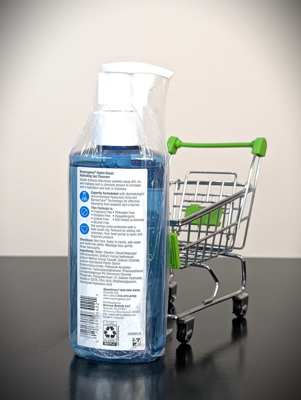 A bottle of Neutrogena Hydro Boost Hydrating Gel Cleanser in blue packaging, wrapped in a clear plastic protective sleeve, standing on a dark surface next to a small metal shopping cart with green handles.