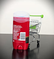 A red deodorant stick with its cap removed stands on a glossy black surface next to a miniature metal shopping cart with a green handle and seat, against a light gray studio background.
